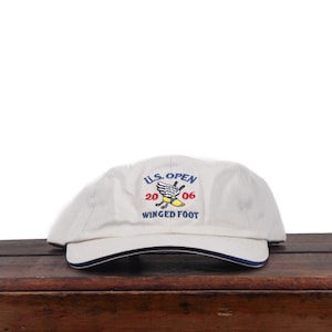 May include: An off-white baseball cap featuring an embroidered U.S. Open 2006 Winged Foot logo on the front. The design includes a winged yellow foot graphic with blue and red text. The cap has a curved brim with a thin dark blue trim along the edge.