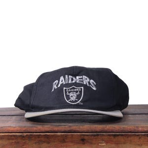 以下が含まれることがあります： グレーのつばが付いた黒いレイダースの野球帽。「Raiders」の文字が、シルバーと白のチームロゴの上に銀糸で刺繍されています。帽子は木製の表面に置かれています。
