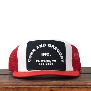 May include: A red, white, and black trucker hat with the text "COHN AND GREGORY INC." on a black patch. The hat has a red brim and mesh back, with the company's location and phone number printed below the logo.