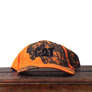 May include: An orange and camouflage baseball cap with the word "CAT" embroidered in black. The cap has a curved brim and a structured crown. The hat is made of a durable material and is suitable for outdoor activities.