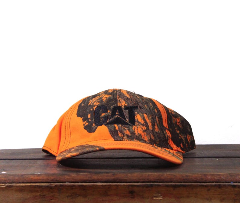 May include: An orange and camouflage baseball cap with the word "CAT" embroidered in black. The cap has a curved brim and a structured crown. The hat is made of a durable material and is suitable for outdoor activities.