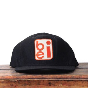 May include: Black baseball cap with a flat peak. The cap has a white square patch with the letters "be" in orange and a small orange dot.