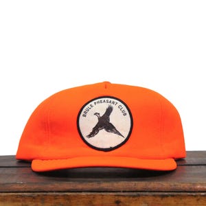 Może przedstawiać: Pomarańczowa czapka myśliwska z białą naszywką przedstawiającą bażanta i napis "BRULE PHEASANT CLUB".