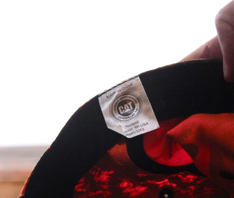 May include: Close-up of a baseball cap's interior, featuring a vibrant orange and black pattern. The cap's lining is a bright orange, and the tag displays the CAT Caterpillar logo and "Made in China".