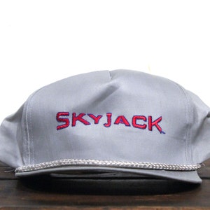 Puede incluir: Una gorra de béisbol gris y blanca con el texto bordado "SKYJACK" en rojo y azul.