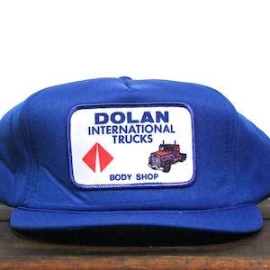 May include: Blue trucker hat with a white patch that says "Dolan International Trucks Body Shop" and features a red diamond and a red semi-truck.
