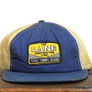 Puede incluir: Gorra de camionero azul y beige con un parche amarillo que dice "The Lane Construction Corp. Steel Tunnel Rehab."