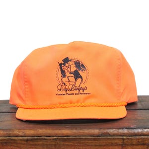 Peut inclure: Une casquette de baseball orange avec un logo noir et blanc qui dit "Lily Langtry's Victorian Theatre and Restaurant".