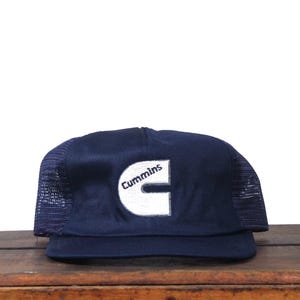 Op de afbeelding: Marineblauwe truckerpet met een wit geborduurd "Cummins" logo. De pet heeft een mesh achterkant en een gebogen klep. De pet ligt op een houten oppervlak.