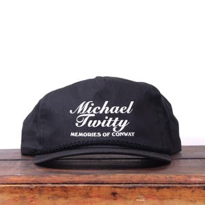 May include: Black cap featuring the white text "Michael Twitty" in a script font, with "MEMORIES OF CONWAY" underneath. The cap has a black rope detail and is displayed on a wooden surface.