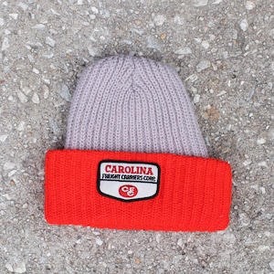 Puede incluir: Un gorro de punto rojo y gris con un parche que dice "CAROLINA FREIGHT CARRIERS CORP." y un logotipo de "CF&C".