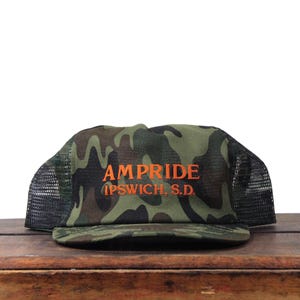 May include: Camouflage print trucker hat with the word "AMPRIDE" in orange lettering. Below the word "AMPRIDE" is the text "IPSWICH S.D." in orange lettering. The hat has a black mesh back and a curved brim.