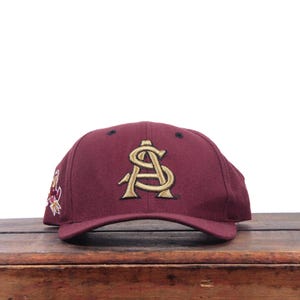May include: A maroon baseball cap featuring a gold embroidered AS monogram on the front panel. The cap has a structured crown, a curved brim, and a small embroidered mascot logo on the side panel. The design includes black eyelets on the crown.