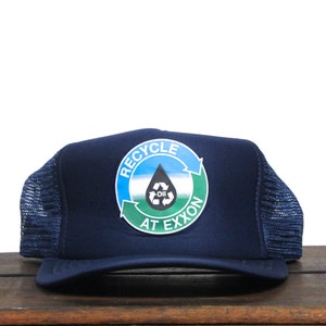 Könnte beinhalten: Eine marineblaue Trucker-Kappe mit einem weißen, grünen und blauen runden Aufnäher, auf dem "Recycle at Exxon" steht, mit einem stilisierten Öltropfen und einem Recycling-Symbol.