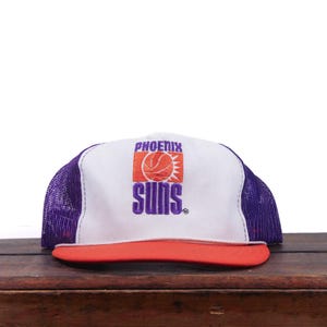 Puede incluir: Una gorra de camionero blanca y morada con visera naranja y el logotipo de baloncesto de los Phoenix Suns bordado en el panel frontal. La gorra tiene una corona frontal blanca, paneles laterales y traseros de malla morada y visera naranja.
