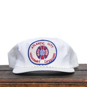 Könnte beinhalten: Weisse Trucker-Cap mit Mesh-Rückseite und Kordelbesatz. Auf der Vorderseite befindet sich ein kreisförmiger Aufnäher mit den Worten "Atlantic City Beach Honorary Lifeguard" in Blau und Rot.