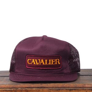 Puede incluir: Gorra de camionero burdeos con parte trasera de malla y panel frontal liso. La parte delantera presenta un parche rectangular con la palabra "CAVALIER" en letras doradas. El sombrero está sobre una superficie de madera.