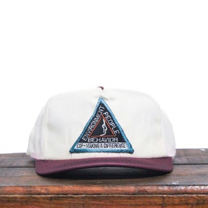 Könnte beinhalten: Eine weiße Baseballkappe mit einem burgunderfarbenen Schirm. Die Vorderseite der Kappe zeigt einen dreieckigen Aufnäher mit den Worten "Umwelt, Menschen, Verhalten, ESP - Einen Unterschied machen".