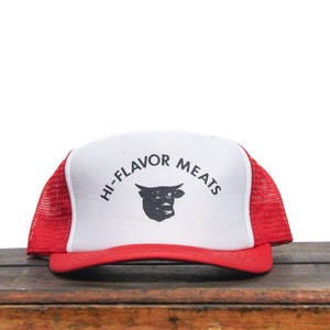 Könnte beinhalten: Eine rot-weiße Trucker-Kappe mit einem schwarzen Bullen-Logo und dem Text "Hi-Flavor Meats" in Schwarz.