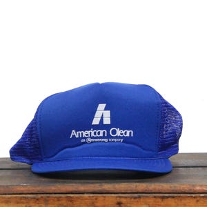 Op de afbeelding: Een blauwe en witte trucker-pet met het logo "American Olean, an Armstrong company".