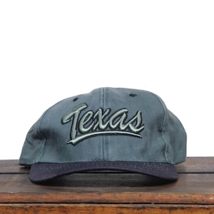 Puede incluir: Gorra de béisbol gris con visera azul oscuro. La palabra "Texas" está bordada en una fuente cursiva en la parte delantera de la gorra en verde y negro.