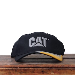 May include: Black baseball cap with the CAT logo in gray and yellow. The hat is sitting on a wooden surface. The brim of the hat is yellow and gray.
