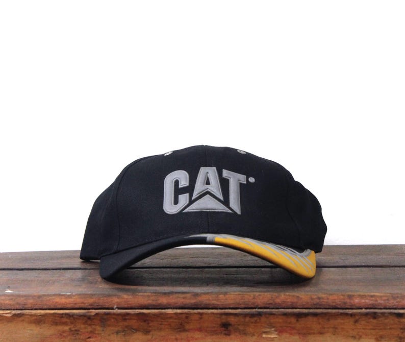 May include: Black baseball cap with the CAT logo in gray and yellow. The hat is sitting on a wooden surface. The brim of the hat is yellow and gray.