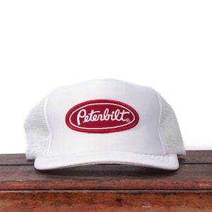 May include: White trucker hat with a red and white Peterbilt logo patch. The hat has a mesh back and a curved peak. The Peterbilt logo is an oval shape with the brand name in a cursive font.