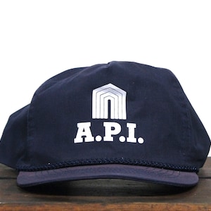 以下が含まれることがあります： 白いロゴにスタイリッシュな建物と「A.P.I.」の文字が書かれたネイビーブルーのベースボールキャップ