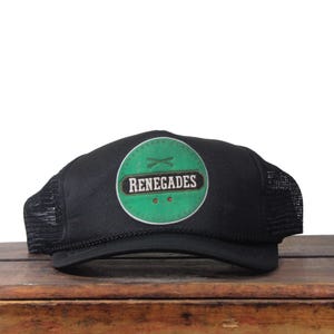 May include: Black trucker hat with a green and white circular patch that reads "RENEGADES". The patch features crossed arrows. The hat has a mesh back and a curved brim.