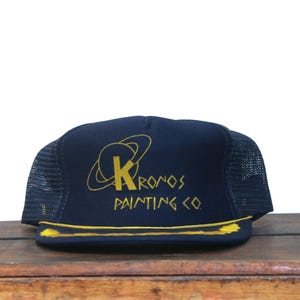 May include: Navy blue trucker hat with a mesh back and a yellow brim. The front of the hat features the text "KRONOS PAINTING CO." in yellow, along with a stylized graphic of a letter "K" inside a circle.