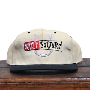 May include: A beige baseball cap with a black brim. The front of the cap features a white horseshoe with the words "MARTY STUART" embroidered in black and red.