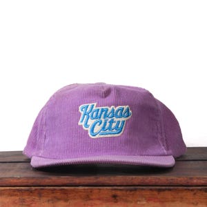 May include: A purple corduroy baseball cap with the embroidered text "Kansas City" in blue and white. The cap is sitting on a wooden surface.