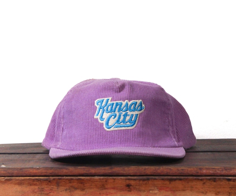May include: A purple corduroy baseball cap with the embroidered text "Kansas City" in blue and white. The cap is sitting on a wooden surface.