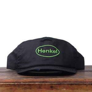 Puede incluir: Gorra de béisbol negra con visera curva. La gorra tiene la palabra "Henkel" bordada en verde dentro de un óvalo verde en la parte delantera. La gorra está sobre una superficie de madera.