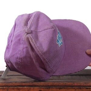 May include: A lavender corduroy baseball cap with a curved brim. The cap has a button on top and a small embroidered design in blue and white on the side. The cap is resting on a wooden surface.