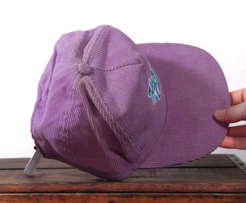 May include: A lavender corduroy baseball cap with a curved brim. The cap has a button on top and a small embroidered design in blue and white on the side. The cap is resting on a wooden surface.