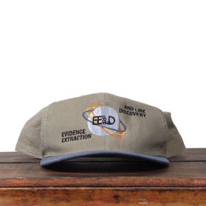 Könnte beinhalten: Khaki Baseball Cap mit blauem Schirm. Die Kappe zeigt den Text "E&D Evidence Extraction" und "And Link Discovery" mit einer Grafik eines Planeten und umlaufenden Ringen.