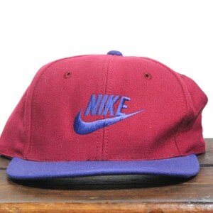 May include: A vintage Nike baseball cap with a burgundy crown and a blue brim. The Nike logo is embroidered in blue on the front of the cap.