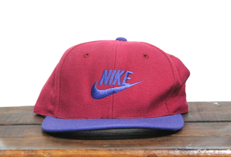 May include: A vintage Nike baseball cap with a burgundy crown and a blue brim. The Nike logo is embroidered in blue on the front of the cap.