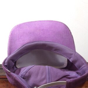 May include: A close-up view of a purple corduroy baseball cap. The cap features a curved brim and a mesh interior for breathability. The adjustable strap is visible at the back. The cap is a solid color.
