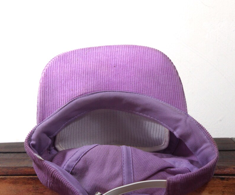 May include: A close-up view of a purple corduroy baseball cap. The cap features a curved brim and a mesh interior for breathability. The adjustable strap is visible at the back. The cap is a solid color.