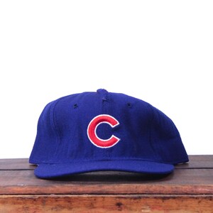 Puede incluir: Gorra de béisbol azul con un logotipo "C" rojo y blanco. La gorra está hecha de un material suave y tiene una visera curva. La gorra está sobre una superficie de madera.