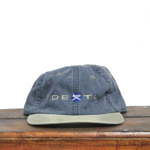 May include: A dark blue and tan baseball cap with the word "DeXTa" embroidered in yellow on the front. The logo features a blue and white X with a yellow outline.