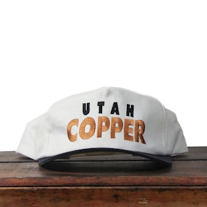 May include: White baseball cap with the words "UTAH COPPER" embroidered in black and copper-colored thread. The cap has a dark blue brim and is displayed on a wooden surface.