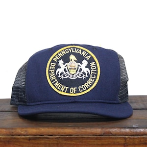 May include: A navy blue trucker hat with a black mesh back. The hat features a yellow and black embroidered patch with the text "Pennsylvania Department of Correction".