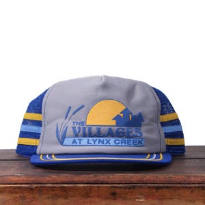 Op de afbeelding: Grijze truckerpet met blauwe en gele details. De voorkant toont de tekst "THE VILLAGES AT LYNX CREEK" met een gele zon en een blauw silhouet van een gebouw. De pet heeft een blauwe mesh achterkant.