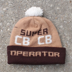 May include: A brown and white knit beanie with a white pom pom on top. The beanie has the text "SUPER CB CB OPERATOR" on it.