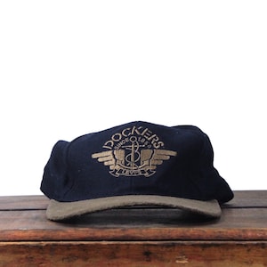 Peut inclure: Casquette Dockers bleu marine avec une visière kaki. Le chapeau présente un texte brodé en or et un logo qui se lit "Dockers Since 1850 Levi's". Le chapeau est posé sur une surface en bois.