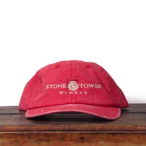 Puede incluir: Gorra de béisbol roja con el texto "STONE TOWER WINERY" bordado en blanco. La gorra tiene una visera curva y está hecha de un tejido suave. Descansa sobre una superficie de madera.
